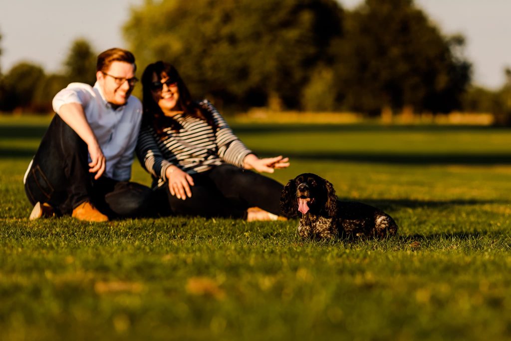 A couple is sitting on the grass in a sunny Leicestershire park, smiling and relaxing. Their small black and brown spaniel is happily sitting in the foreground, making this serene pre-wedding moment picture-perfect. Trees are blurred in the background.