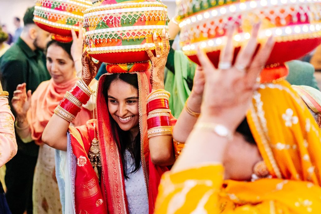 A group of people in colourful traditional attire participate in a vibrant cultural event, reminiscent of a traditional Sikh pre-wedding celebration in London. Women gracefully carry decorated pots on their heads, adding to the joyful atmosphere that highlights rich cultural heritage.