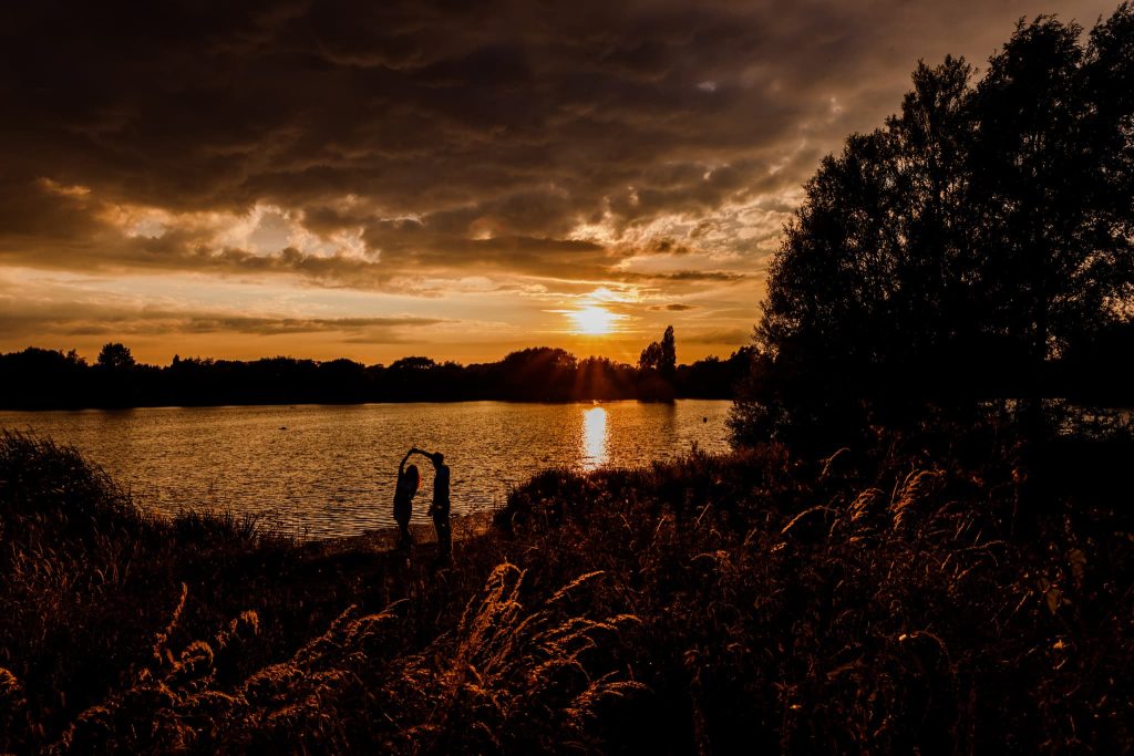 A couple stands by the lake at Watermead Park under a dramatic sunset, with dark clouds filling the sky and the sun casting a warm glow across the water. Tall grasses and trees are silhouetted against the vibrant sky, creating an enchanting scene perfect for their pre-wedding moment.