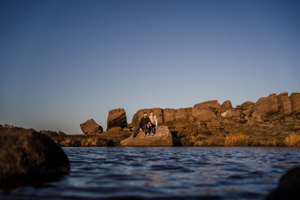 A couple sits on a large rock surrounded by water, capturing their pre-wedding bliss in the serene beauty of the Peak District, with a rocky landscape and clear blue sky completing the picturesque backdrop.