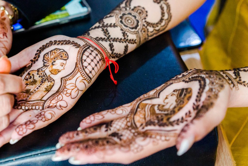 Two hands with intricate henna designs celebrate Indian tradition. One hand showcases a detailed portrait of a person in traditional attire, while the other is adorned with elaborate patterns and floral motifs. Both hands rest gracefully on a black surface, exuding cultural richness.