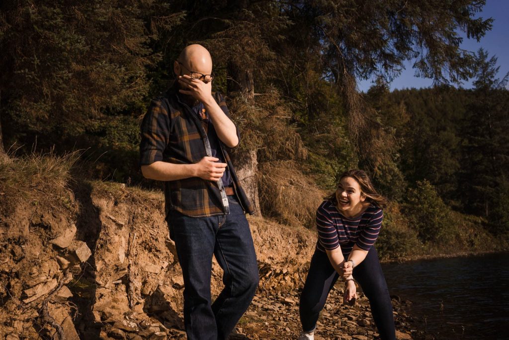 A man in a plaid shirt covers his eyes while a woman in a striped sweater laughs with her hands together. They stand on a rocky path by a forested lake, enjoying the sunny day—which sets the perfect scene for their Peak District pre-wedding adventure.