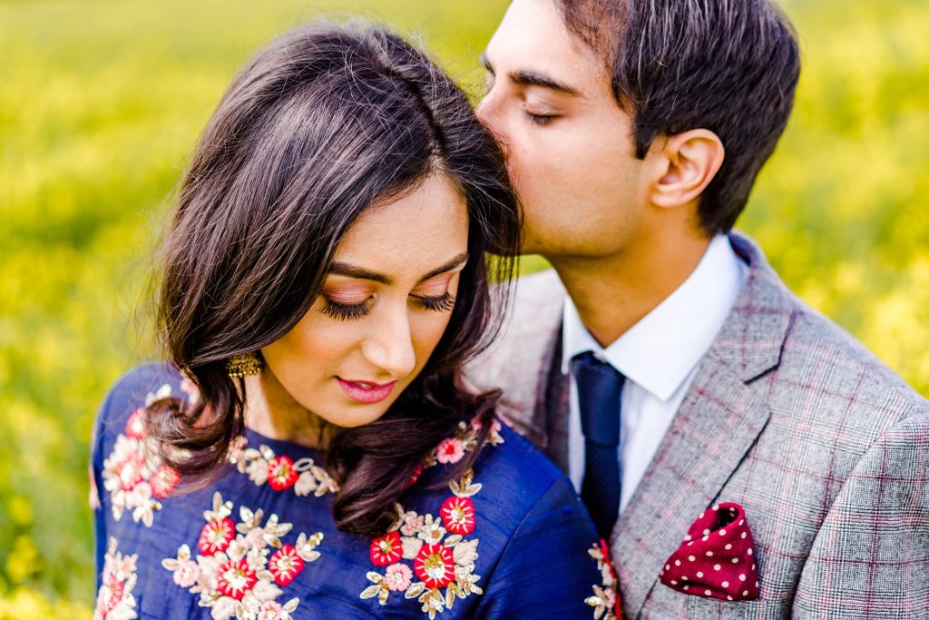 In this serene couple photography shot, a man gently kisses the womans forehead as she smiles softly in her floral embroidered dress. He dons a plaid suit with a dotted pocket square, all set against a backdrop of vibrant greenery.