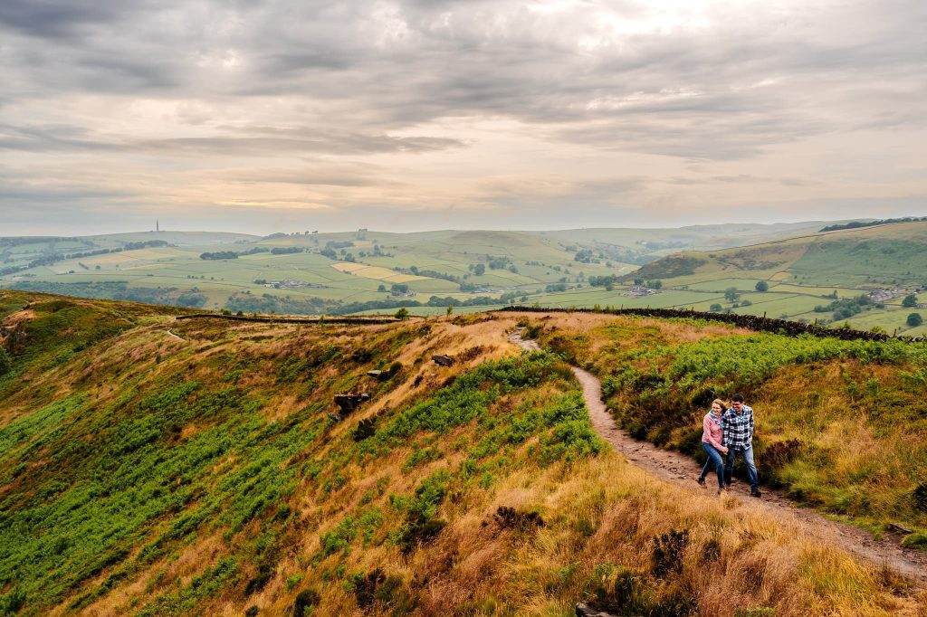 A couple enjoys a pre-wedding stroll along a winding dirt path on a grassy hillside in the Peak District, with lush green fields and small villages unfolding in the background. The overcast sky adds to the serene and picturesque landscape.