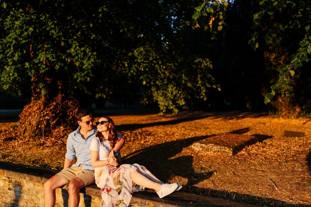 A couple sits on a stone wall in an Oakham park, enjoying a sunlit, pre-wedding afternoon. The woman, in a floral dress and sunglasses, leans against the man in a striped shirt and shorts. Trees and golden leaves create a serene autumn backdrop.