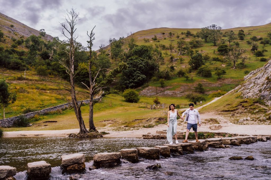 A couple steps across stone blocks over a river in the scenic landscape of Dovedale, with lush hills and sparse trees under a cloudy sky. The woman, in white, and the man, in a t-shirt and shorts, enjoy their pre-wedding moment. A lone tree stands nearby.