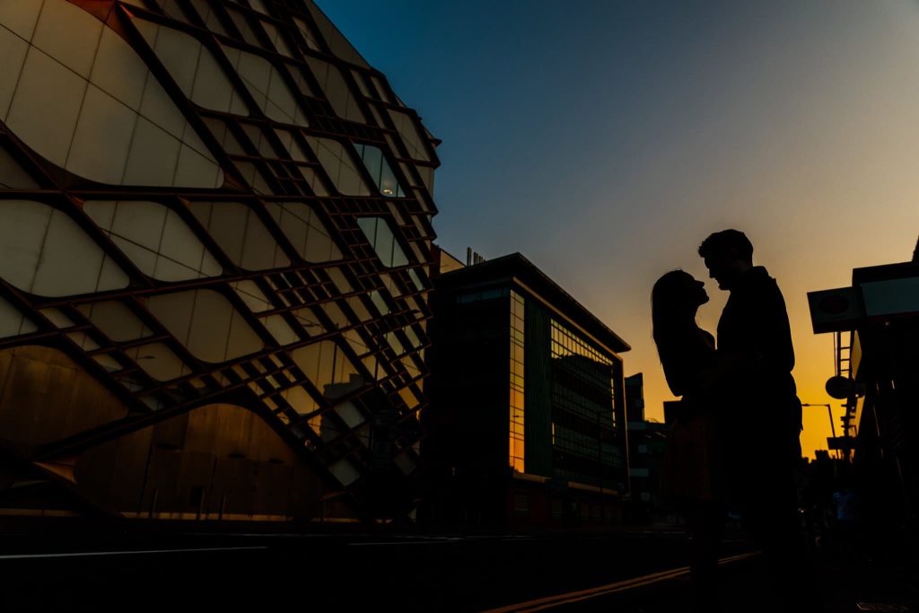 Silhouette of a couple embracing in front of Sheffield's modern buildings with intricate patterns, under a vibrant sunset sky, capturing the magic of their pre-wedding moment.