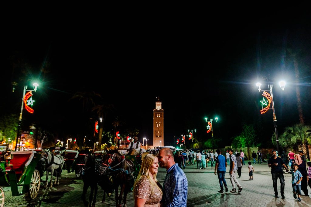 A couple stands smiling at each other in a lively Marrakesh square filled with people, horse-drawn carriages, and illuminated street lamps, capturing the magic of their pre-wedding moment. A tall tower is visible in the background under the night sky.