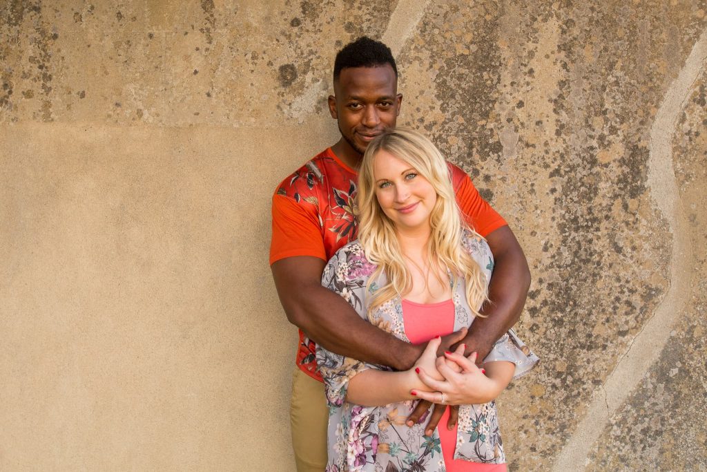 A couple stands against a textured wall at Prestwold Hall, smiling. The man, in a red patterned shirt, embraces the woman from behind. She wears a floral top over a pink shirt. They look content and relaxed as they enjoy their pre-wedding moment together.
