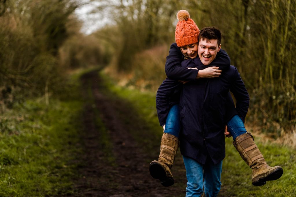 On a woodland path in Lincolnshire, a person wearing an orange knit hat playfully gives a piggyback ride to another. Both are all smiles, dressed in casual outdoor attire, embodying the joy of a pre-wedding shoot amidst towering trees and rustic trails.