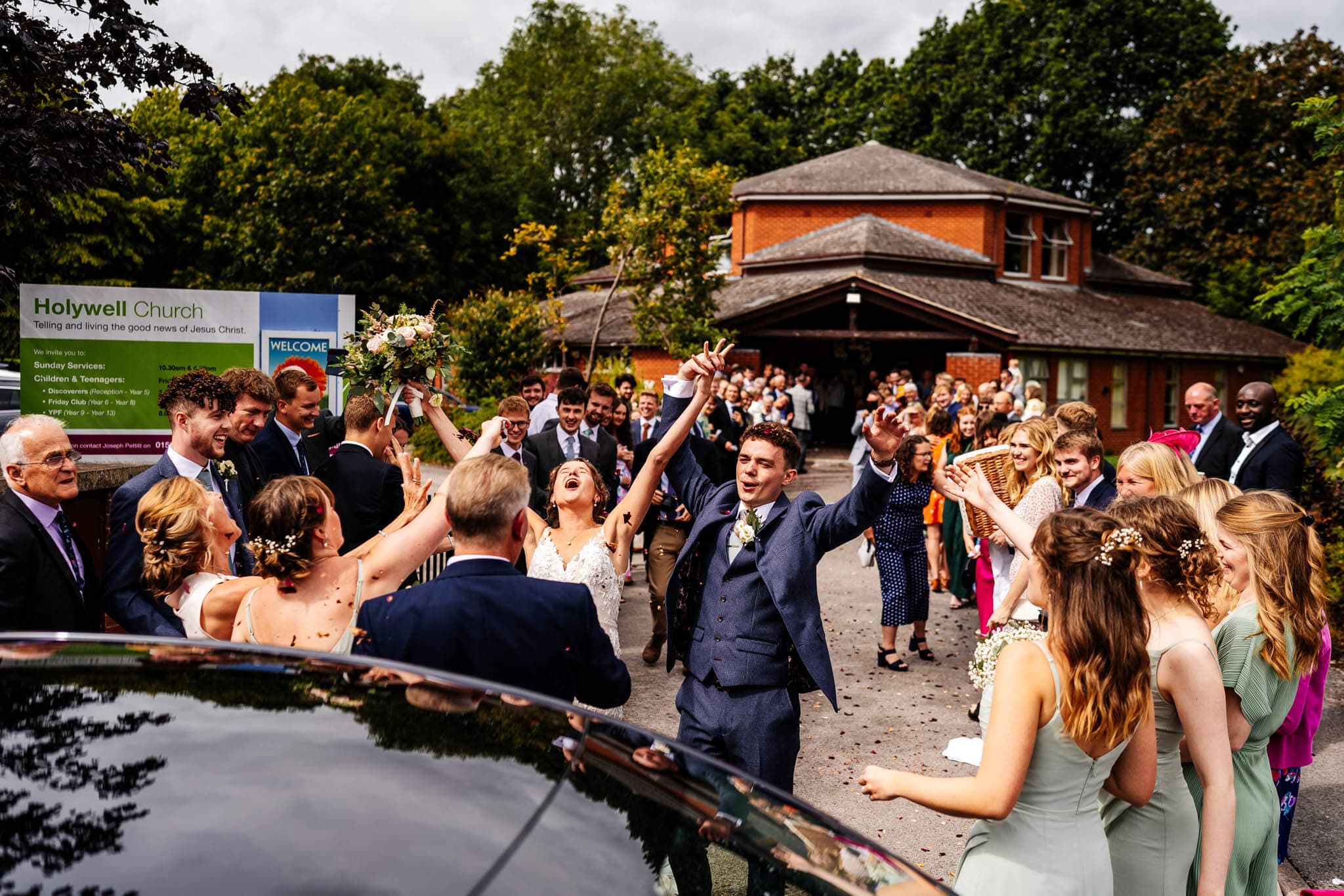 bride and groom raise their hands in happiness at the end of a very busy confetti run outside of their Leicestershire church wedding.