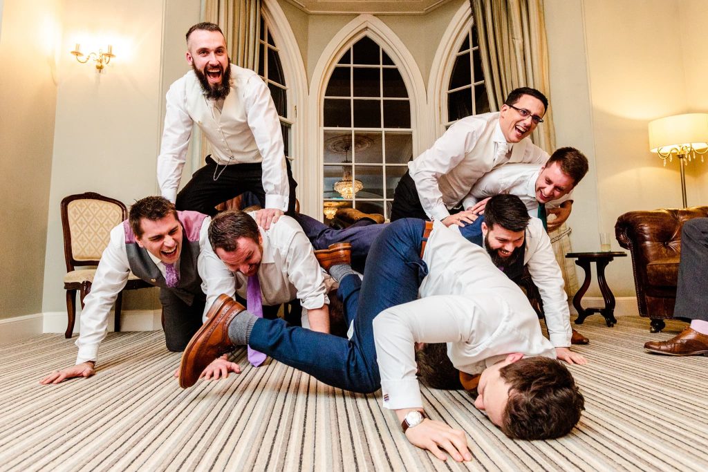 A group of seven men in formal attire joyfully form a human pyramid indoors. One man is on the floor, supporting others stacked on top of him. They appear to be laughing and enjoying the moment in a room with large windows and elegant decor. fun candid Warwick House wedding in Warwickshire