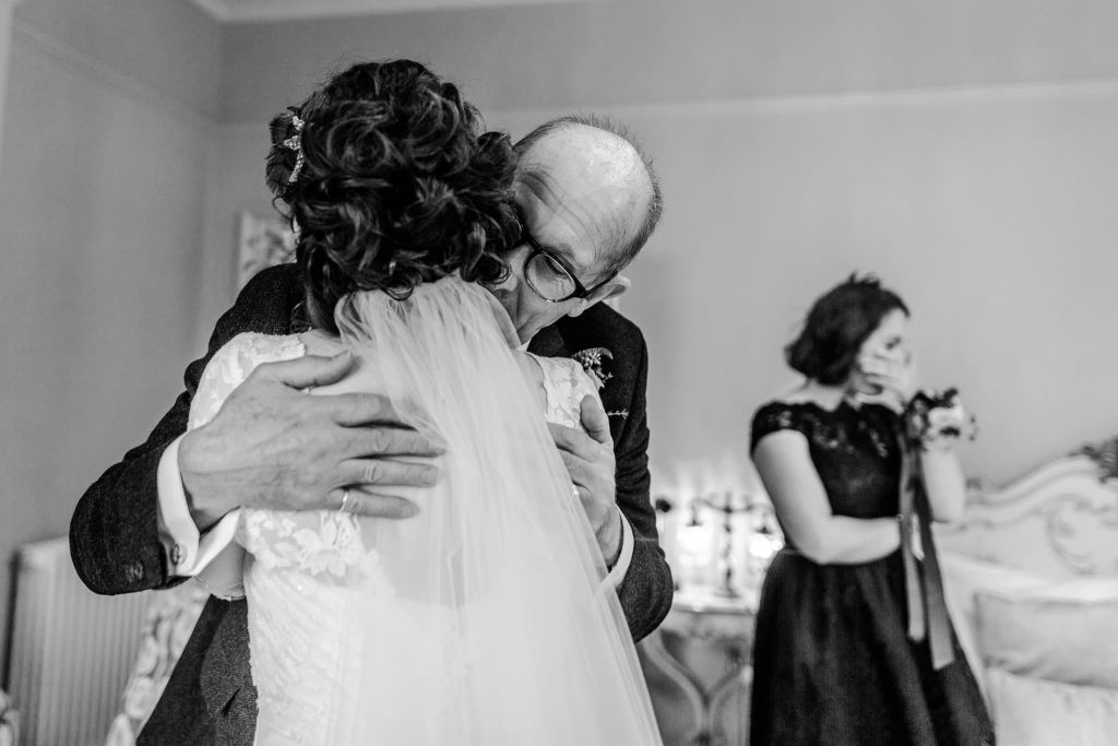 A black and white photo captures the emotional embrace of a father hugging his daughter the bride, her lace dress and veil adding elegance. In the background, during the first look, a woman in a dark dress dabs her eyes with a handkerchief, clearly moved by the touching father of the bride moment.