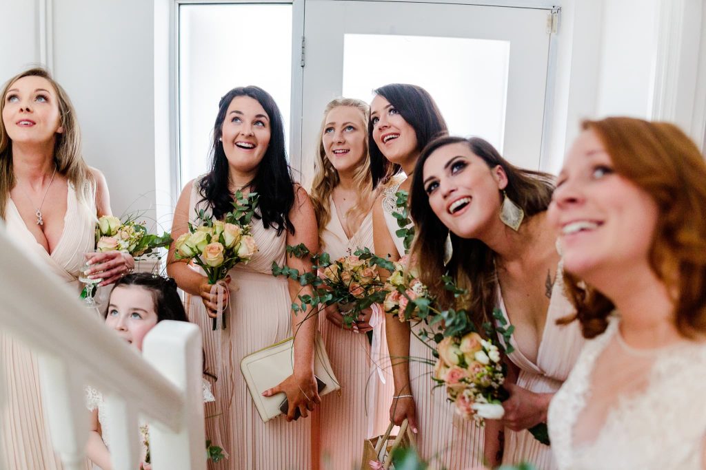 A group of six bridesmaids in matching light pink dresses and one young girl holding bouquets of flowers look up excitedly inside a bright room with a windowed door during their first look, capturing the cheerful anticipation of wedding preparations.