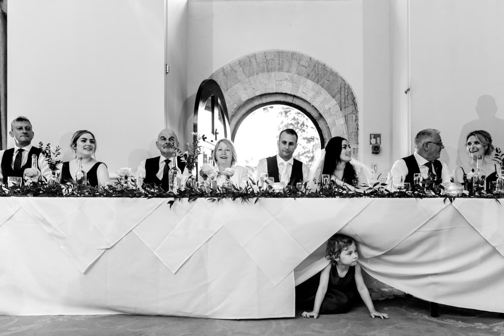 In a black and white photo at Stapleford Park, a wedding party sits elegantly at a long, decorated table. A small child playfully peeks out from under the white tablecloth, adding charm to the formal gathering.