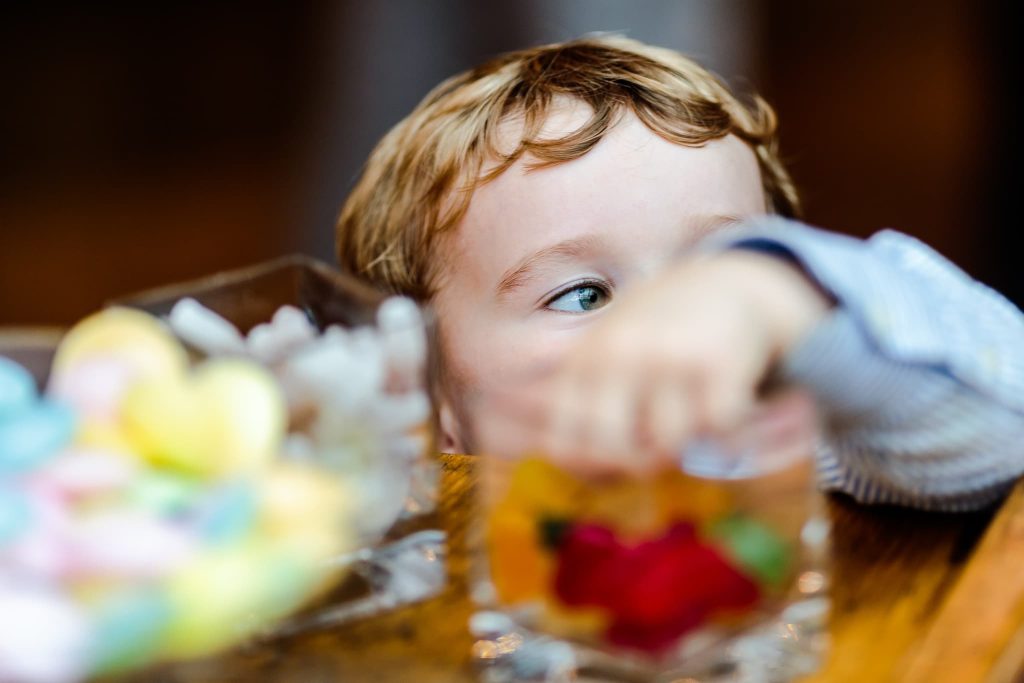 A young child with light brown hair peeks over the edge of a wooden table at a wedding, reaching for a clear bowl filled with sweets, their curious eyes and outstretched hand capturing the whimsical spirit of kids at weddings.