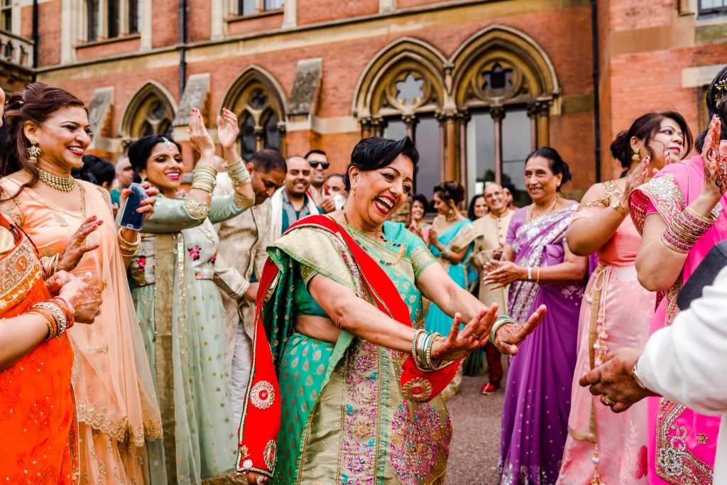 A group of people in colourful traditional attire joyfully dance together outside Kelham Halls arched windows. Celebrating a Hindu wedding, they smile and clap, creating a festive and lively atmosphere.
