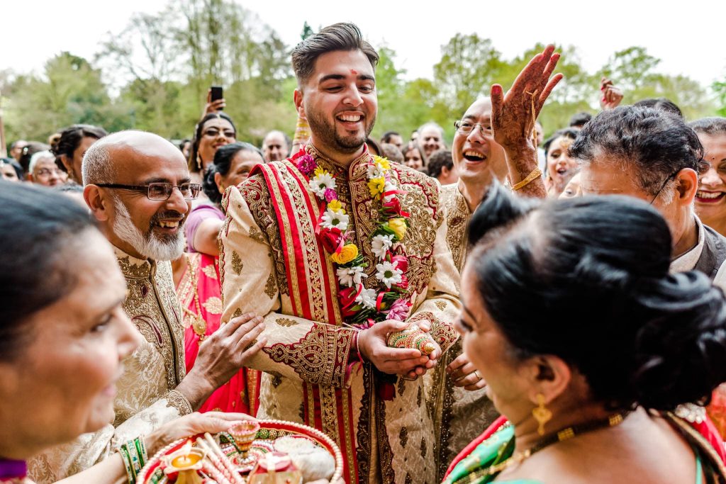 A groom, making a grand entrance in line with Hindu traditions, is dressed in a traditional outfit adorned with a floral garland. He smiles joyfully amidst the lively outdoor wedding celebration, surrounded by cheerful people, some holding trays with ceremonial items.