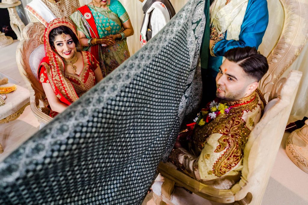 A couple in traditional Indian wedding attire sits on ornate chairs, separated by a decorative mesh screen during a Hindu ceremony. The bride dazzles in a red and gold saree, while the groom dons a gold sherwani. Nearby, guests in traditional clothing celebrate their union in Nottinghamshire.