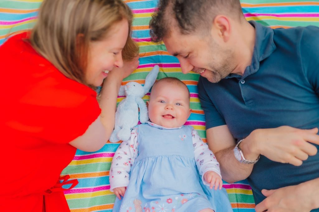 A smiling baby in a blue dress lies on a colorful striped blanket, flanked by a woman in a red top and a man in a blue shirt, both beaming at their little one. The woman holds a plush toy rabbit. Captured by Milton Keynes family photographer, this moment radiates warmth and love.