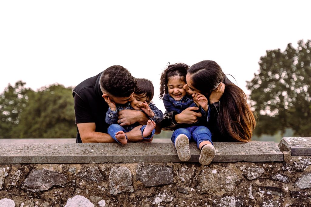 A family of four sits on a stone wall, captured beautifully by a Leicester family photographer. The parents playfully tickle their two young children, sparking bouts of laughter. Casually dressed, they are enveloped by lush greenery in the background.