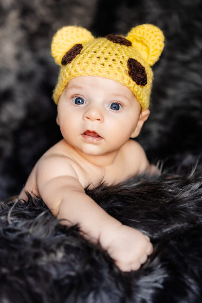A baby with bright blue eyes, captured by a Leicester family photographer, wears a crocheted yellow hat with bear ear spots. The infant lies on a soft, dark surface, gazing at the camera with curiosity.