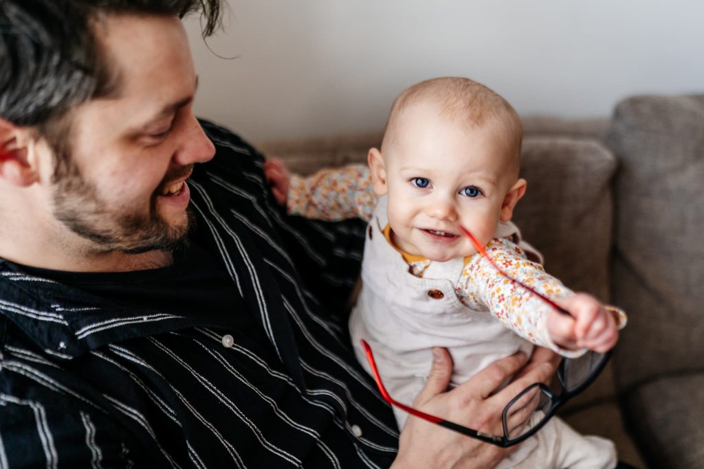 A man smiles while holding a baby on a couch, captured by a Nottingham family photographer. The baby, wearing a patterned shirt and white overalls, reaches towards the camera with a cheerful expression, holding glasses in one hand. The scene is warm and playful.