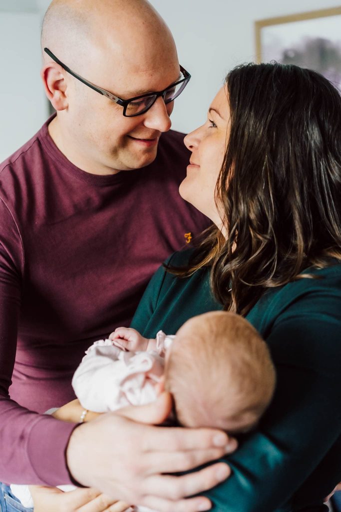 A man and woman smile at each other while holding a sleeping baby wrapped in a pink blanket. Captured by a Leicester family photographer, they appear happy and affectionate, standing indoors with soft lighting.