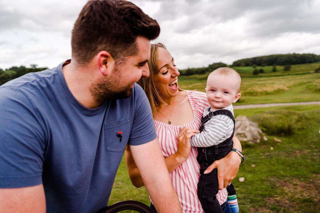 A smiling couple enjoys a moment outdoors captured by a Leicester family photographer. The man in a blue shirt leans towards the woman in a striped top holding the baby, who is wearing a striped shirt and dark overalls. Green fields and a cloudy sky form the picturesque background.