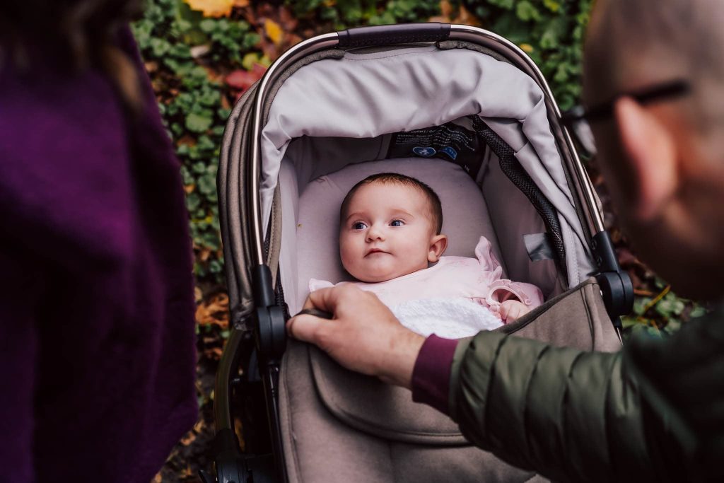 A baby in a stroller looks up at an adult gently touching the stroller, capturing a tender moment perfect for a Leicester family photographer. The baby, dressed in light pink, is surrounded by lush green foliage and fallen leaves in this enchanting outdoor scene.