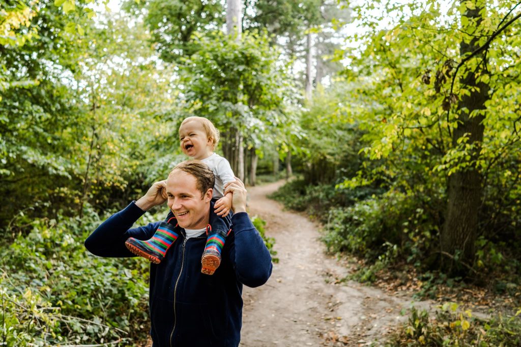 A man strolls along a forest path with a smiling child perched on his shoulders, colorful striped socks brightening the scene. The lush green trees frame this joyous moment, perfect for capturing by a Leicester family photographer.
