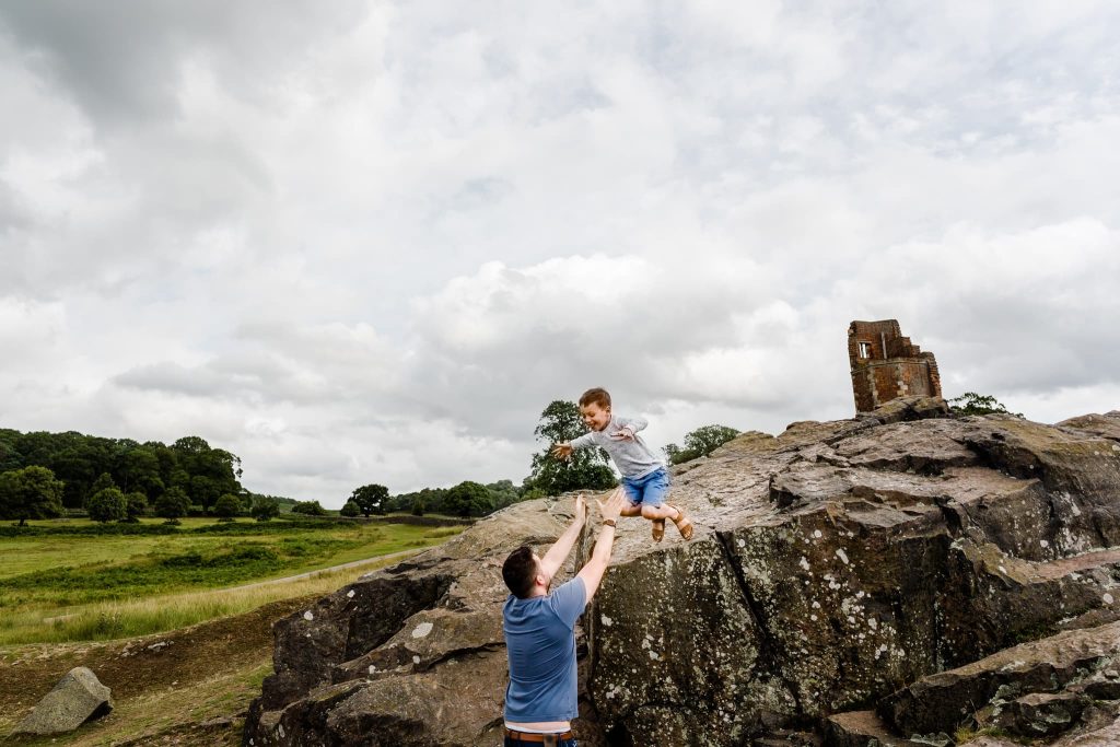 A man on rocky terrain lifts a young boy jumping toward him, captured beautifully by a Leicester family photographer. The scenes backdrop showcases grassy fields, trees, and an old stone structure beneath a cloudy sky.