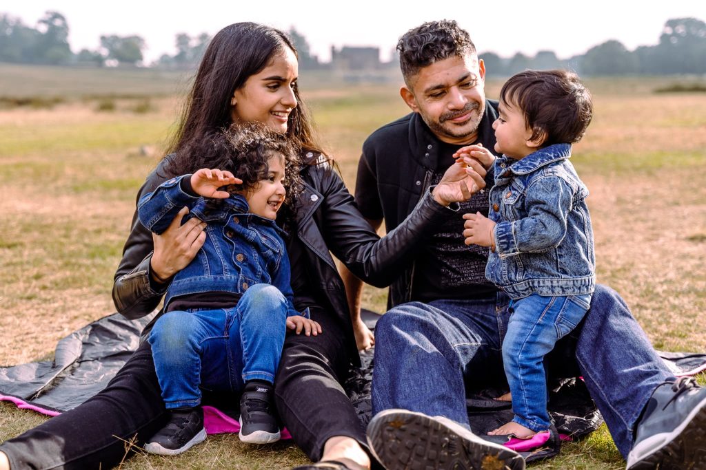 A Leicester family photographer captures a tender moment as a family sits on a blanket in a grassy field. With trees dotting the distant background, the couple smiles at their two young children, one sitting and one standing, all dressed in cozy jeans and jackets.