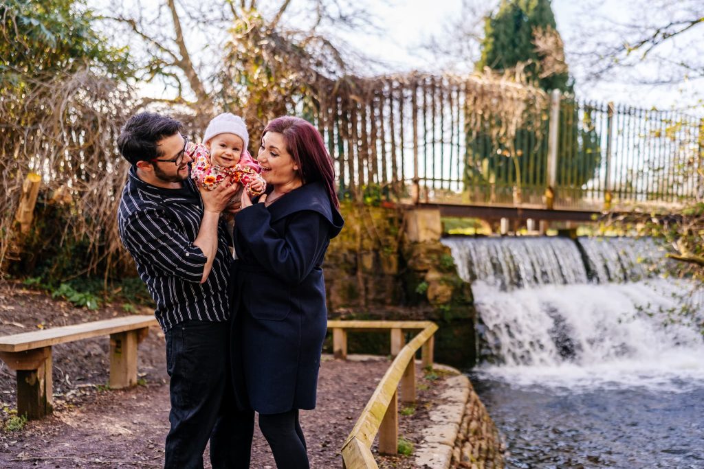 A family of three stands near a small waterfall in a park, captured by a Nottingham family photographer. The father holds a smiling baby dressed in colorful clothes, while the mother stands beside them, her eyes lovingly on the baby. A wooden fence and trees frame this delightful scene.