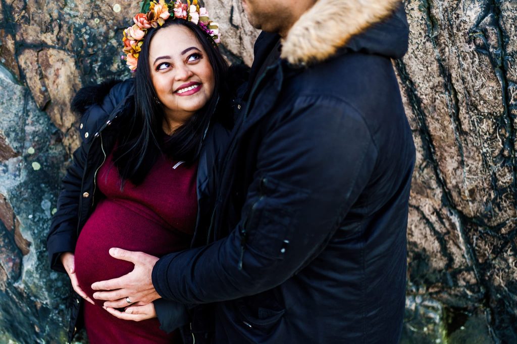 A pregnant woman wearing a flower crown and a burgundy dress smiles up at a man beside her. The Leicester maternity photographer captures the moment as the man, in a dark coat with a fur-lined hood, gently places his hands on her belly against a textured rock wall backdrop.