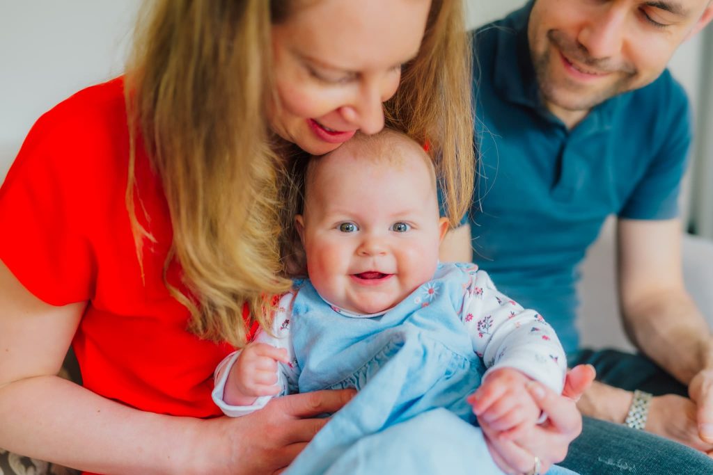 A happy baby in a blue dress sits on a womans lap, smiling at the camera. With the woman in a red top holding the baby and a man in a blue shirt beside her, this delightful scene captured by a Milton Keynes family photographer creates an inviting and warm family moment indoors.