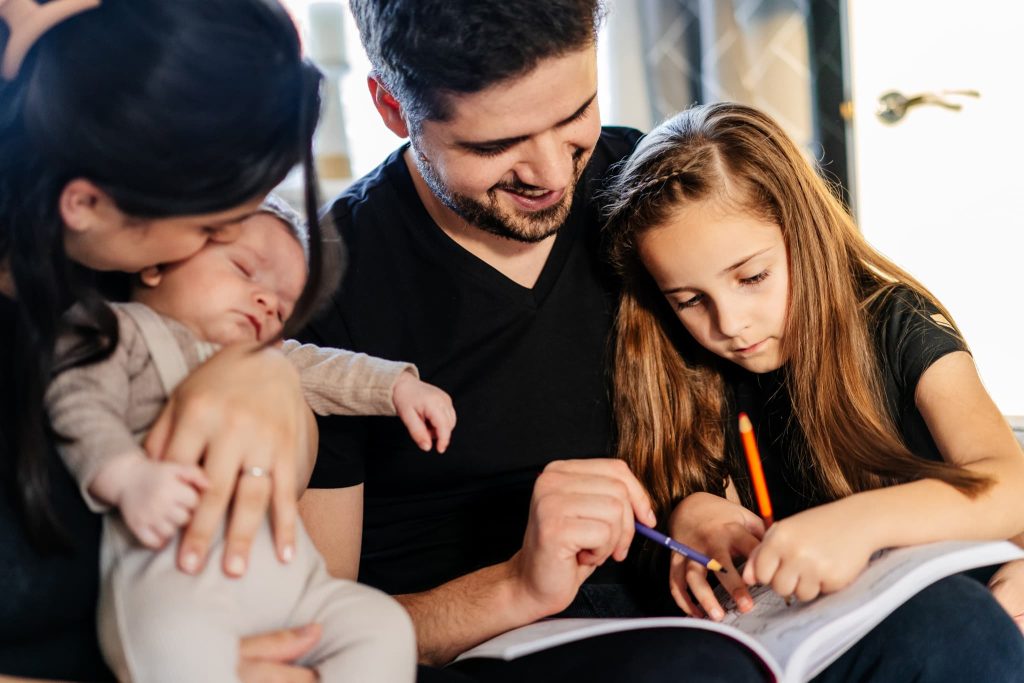 A family sits together, with a mother holding a sleeping baby on her lap, while the father helps a young girl with her homework. Captured by the keen eye of a Leicester family photographer, they are sitting closely, sharing a moment in a warmly lit room.
