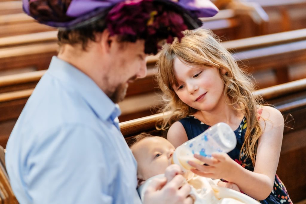 A young girl with long hair lovingly feeds a baby with a bottle while sitting in a church pew, captured beautifully by the Loughborough Christening photographer. A man wearing a large, decorative hat looks on with a gentle smile in this warm and intimate setting.