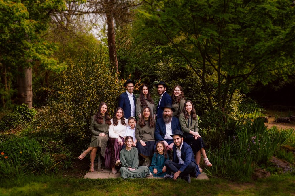 A family of thirteen, captured by a skilled Leicester family photographer, poses outdoors in a lush garden. Dressed in formal attire, they are surrounded by greenery and trees, with some members sitting and others standing around a stone pathway.