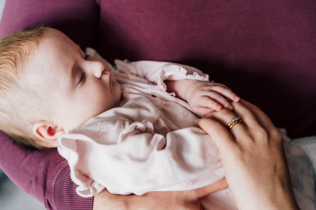 A sleeping baby in a pink outfit is gently cradled in someones arms, captured beautifully by a Leicester family photographer. A hand with a gold ring rests on the babys chest, providing comfort and warmth, evoking tenderness and care.