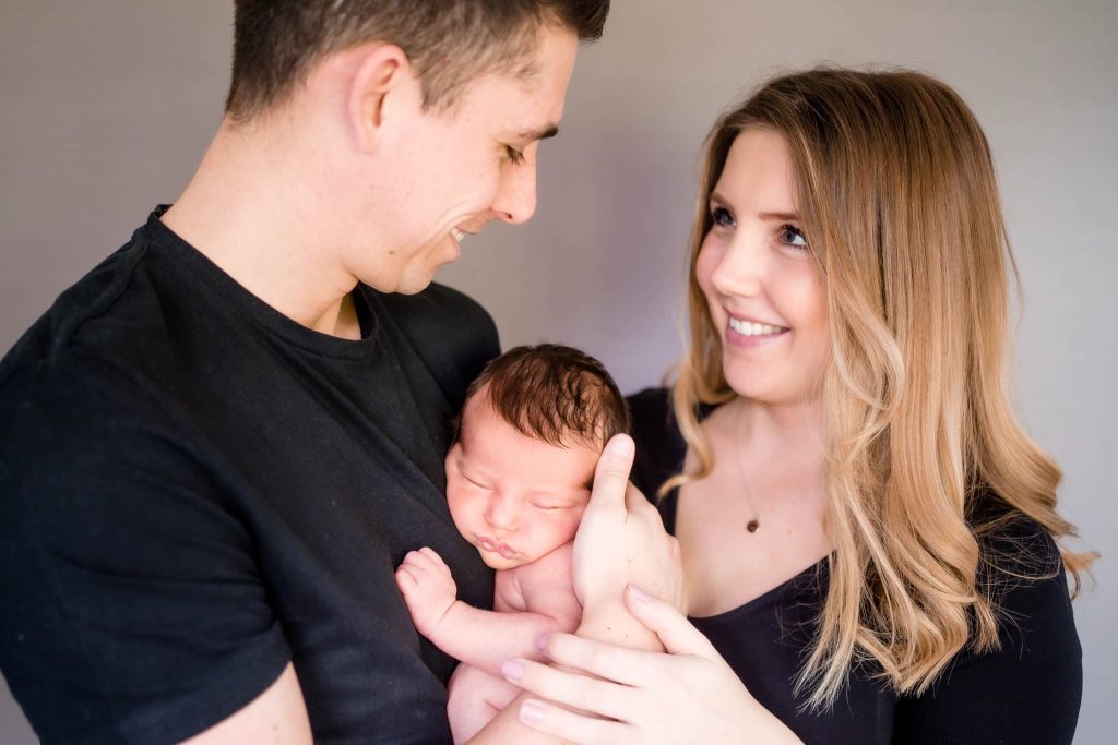 A couple smiling at each other while holding a sleeping newborn, captured perfectly by a Leicester family photographer. The man cradles the baby as the woman gently touches him. They are dressed in black against a plain background, their love and joy perfectly framed in this precious moment.
