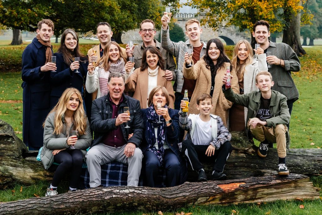 A group of people, including men, women, and children, are gathered outdoors in a park-like setting captured by a Derbyshire family photographer. They are smiling and holding drinks while posing on and around logs surrounded by green grass and the vibrant hues of autumn foliage.
