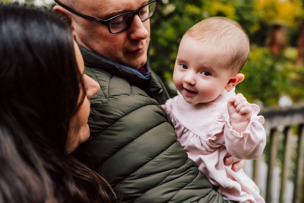 A man in glasses holds a smiling baby dressed in pink, while a woman with long hair looks on. Captured by a Leicester family photographer, they are outdoors with blurred greenery in the background, conveying a warm family moment.