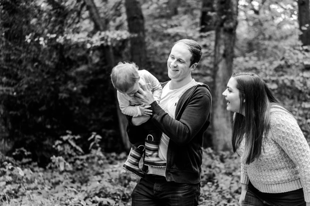 A joyful black and white photo captured by a Leicester family photographer shows two adults and a child in a forest. The man playfully lifts the child, while the woman smiles warmly at them, with trees and lush foliage forming an enchanting background.
