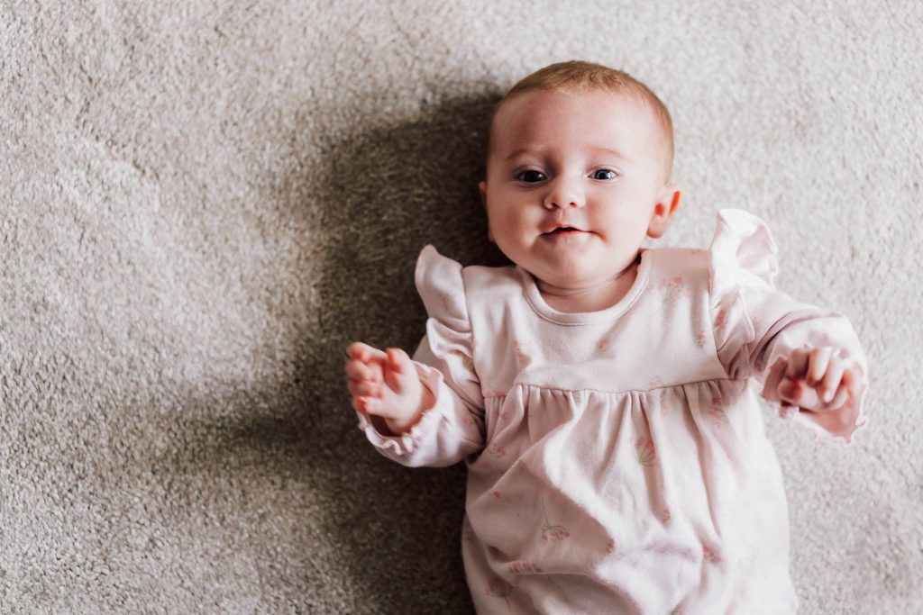 A baby in a light pink outfit with ruffled shoulders lies on a soft, gray carpet. Captured by a Leicester family photographer, the baby gazes upward with a gentle expression, one arm slightly raised.