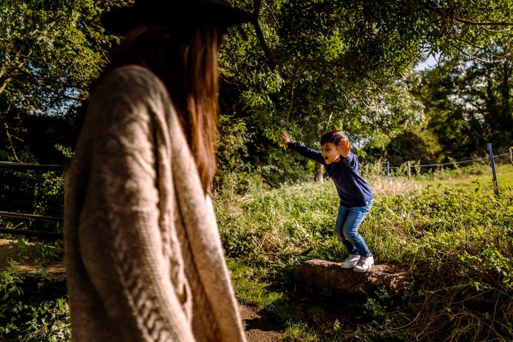 A young child in a blue sweater and jeans jumps off a rock in a grassy, sunlit field. An adult in a wide-brimmed hat and beige sweater watches nearby, capturing the moment like a Leicester family photographer amidst trees and greenery under a clear sky.