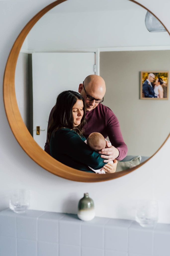 A couple, captured by a Leicester family photographer, is reflected in a circular mirror, tenderly holding their newborn. They stand in a softly lit room with a wall photo of another couple behind them. A small vase and two glasses sit under the mirror, completing the serene scene.
