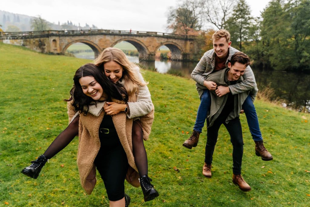 Two siblings give piggyback rides to two siblings on a grassy field near a river, capturing a moment of joy. An old stone bridge and lush trees provide the perfect backdrop for this delightful scene, all skillfully framed by the lens of a Derbyshire family photographer on an overcast day.