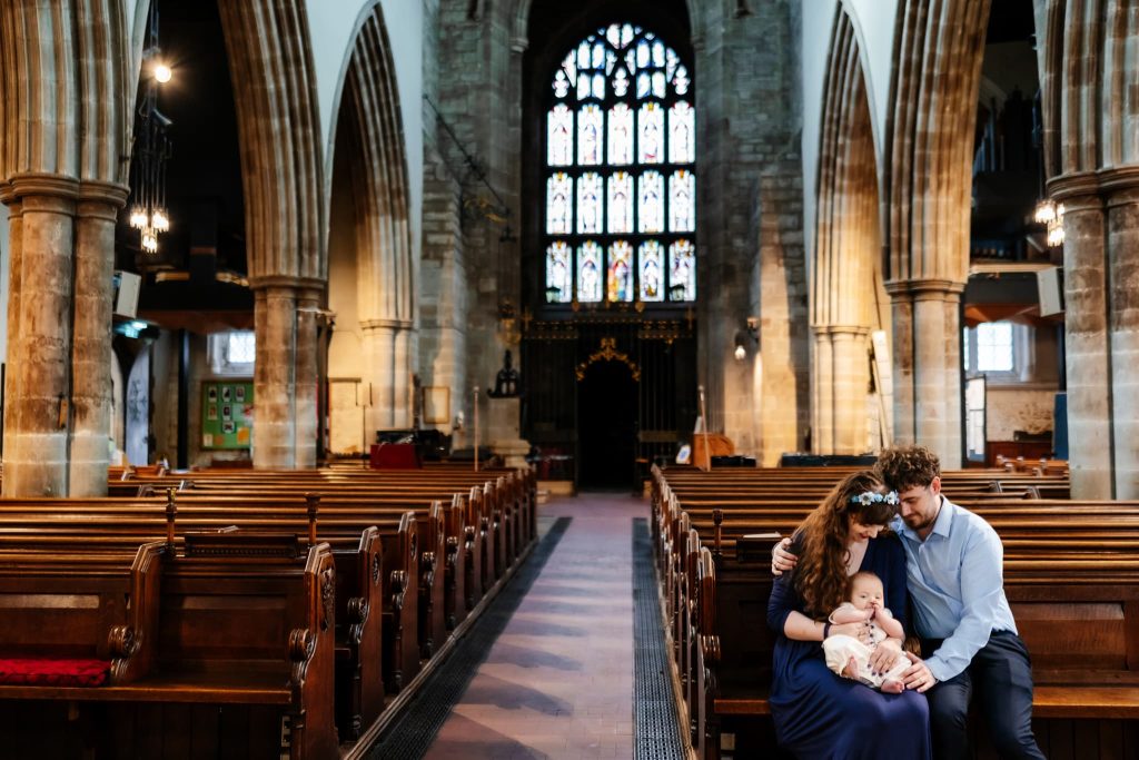 A young couple sits on a pew in a large, ornate church, holding their baby under the soft gaze of a Loughborough Christening photographer. They are surrounded by tall stone arches and stained glass windows, creating a serene and peaceful atmosphere.
