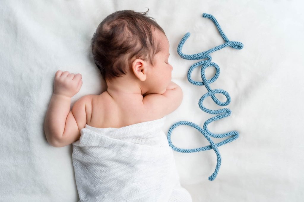 A baby wrapped in a white blanket sleeps on a soft, white surface. Next to the baby, the name Joey is spelled out in blue knitted yarn, captured effortlessly by a Leicester family photographer.